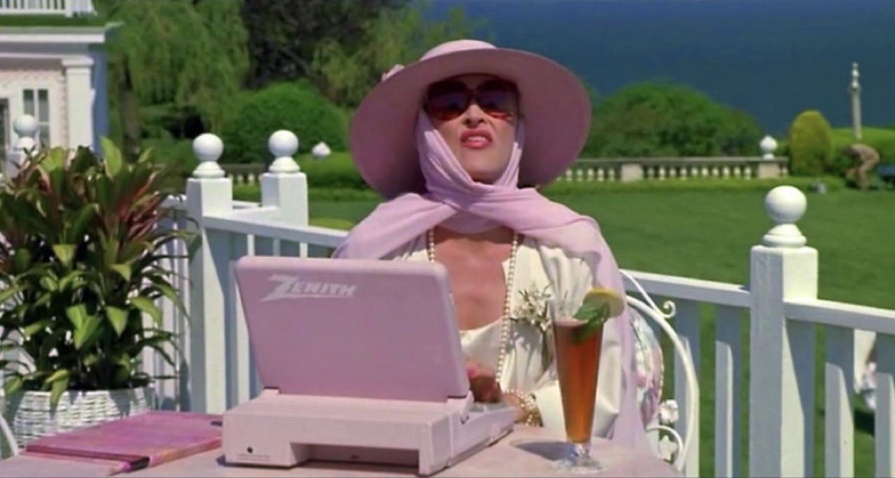 romy & michele scene — woman in pastel sunhat, sunglasses, scarf and pearls at a glass-top patio table with a lavender zenith laptop and an iced tea